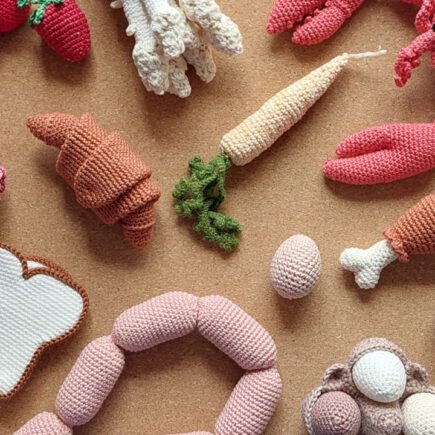 Op een kurk achtergrond liggen diverse gehaakte etenswaren zoals een sneetje wit brood, eieren, stokbrood, slinger worst, kippenbout, kreeft, asperges, aardbeien, croissant, wortel en salami